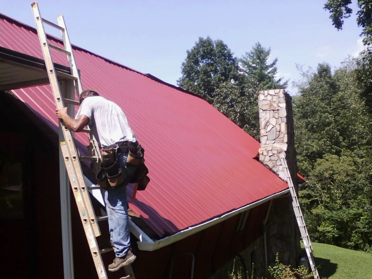 Roofer on a ladder beside a red metal roof and stone chimney — Soffit & Fascia Repair by Roofing Brothers in Hallandale